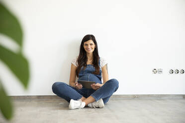 Lächelnde schwangere Frau, die ein digitales Tablet benutzt, während sie an einer weißen Wand in ihrem neuen Haus sitzt - MJFKF00575