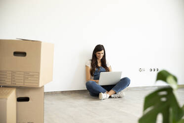 Schwangere Frau benutzt Laptop, während sie auf dem Boden vor einer weißen Wand in einem neuen Haus sitzt - MJFKF00577