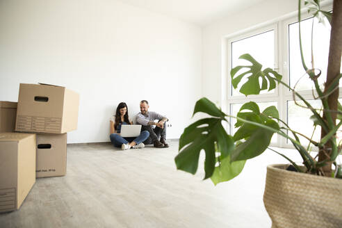 Man sitting with wife using laptop on hardwood floor in new unfurnished house - MJFKF00578