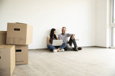 Frau im Gespräch mit ihrem Mann, während sie einen Laptop an der Wand ihres neuen Hauses benutzt - MJFKF00579
