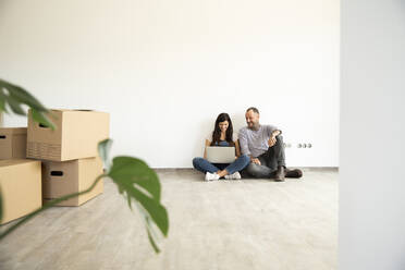 Man sitting with wife using laptop on hardwood floor against wall in new unfurnished home - MJFKF00580