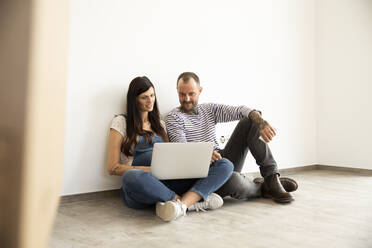 Ein Paar benutzt einen Laptop und sitzt an einer weißen Wand in einer neuen, unmöblierten Wohnung - MJFKF00581
