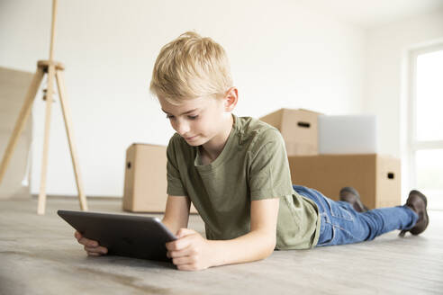 Junge mit blondem Haar, der ein digitales Tablet benutzt, während er in einem neuen Haus auf dem Boden liegt - MJFKF00582