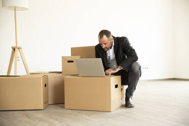 Businessman using laptop on cardboard box while crouching on flooring in new unfurnished home - MJFKF00585