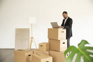 Businessman using laptop on cardboard box while standing in new unfurnished house - MJFKF00586