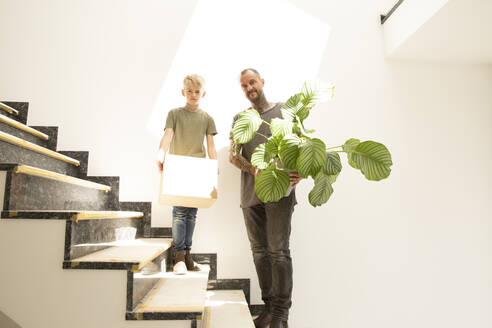 Vater mit Calathea und Sohn mit Kiste auf der Treppe vor der Wand im neuen Haus stehend - MJFKF00602