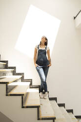 Schwangere Frau mit den Händen in den Taschen, die auf einer Treppe an der Wand eines neuen Hauses steht - MJFKF00603