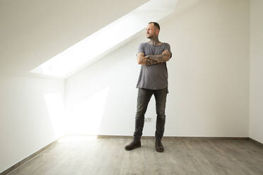 Thoughtful man with arms crossed looking through window while standing in attic of new house - MJFKF00619