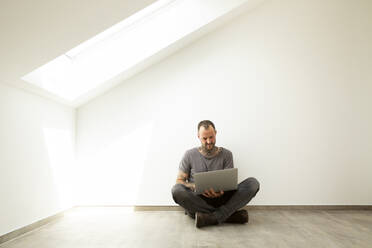 Älterer Mann, der einen Laptop benutzt, während er gegen eine weiße Wand im Dachgeschoss eines neuen Hauses sitzt - MJFKF00620