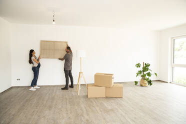 Couple carrying window frame while standing against wall in new unfurnished house - MJFKF00622