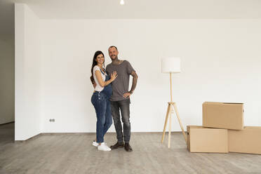 Smiling couple with electric lamp and boxes standing against wall in new house - MJFKF00625