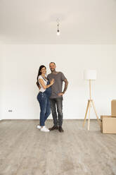 Smiling couple with electric lamp standing against wall in new unfurnished house - MJFKF00626