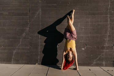 Young woman with red hair performing handstand against black wall - TCEF00983