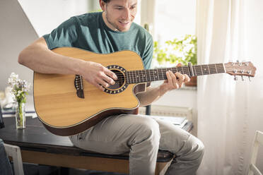 Smiling man playing guitar while sitting on table at home - MSUF00291