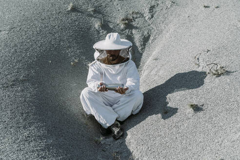 Mann in Imkerkleid sitzt in einer trockenen apokalyptischen Landschaft und hält eine Thermoskanne - AFVF07085