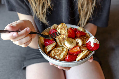 Mid section of teenage girl eating bowl of mini pancakes with strawberries and bananas - SARF04621
