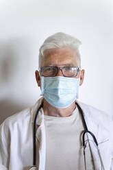 Senior doctor wearing surgical mask on face standing against wall - AFVF07118