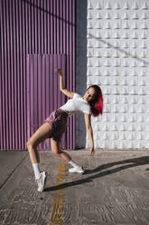 Junge Frau mit gefärbten roten Haaren tanzt vor einer lila Wand in der Stadt - TCEF01019