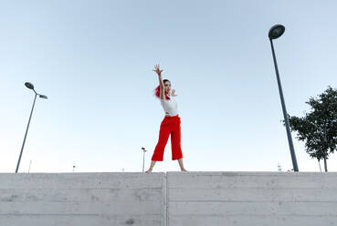 Woman wearing red pant dancing on wall against clear sky - TCEF01026