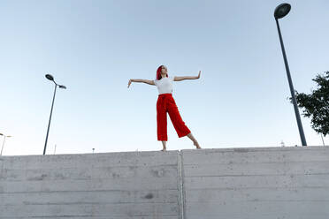 Young dancer with arms outstretched standing on surrounding wall against clear sky - TCEF01028