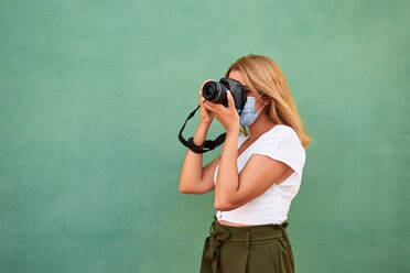 Young woman with a mask and a camera on a green background - CAVF88614