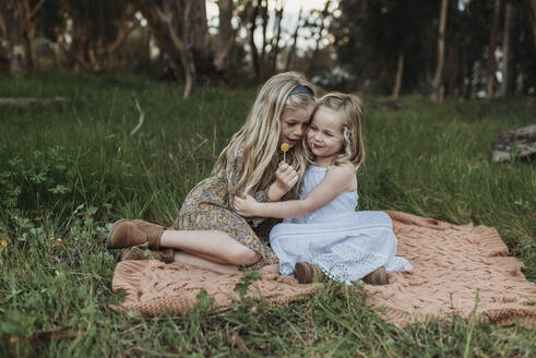 Zwei blonde Schwestern sitzen auf einer Decke in einem Feld mit Lutschern - CAVF88622