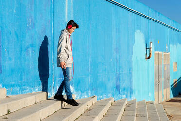 Junger Teenager hört Musik mit Kopfhörern an einer blauen Wand - CAVF88643