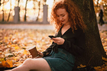 Glückliche junge Frau, die ihr Smartphone benutzt, sitzt im Herbst an einem Baum im Park - CAVF88682
