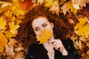 Junge Frau mit Blatt im Gesicht in orangefarbenen Blättern im Herbstpark liegend - CAVF88683