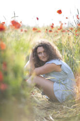 Beautiful woman with hand in hair sitting in poppy field - ASCF01457