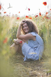 Smiling young woman with hand in hair sitting in poppy field - ASCF01469