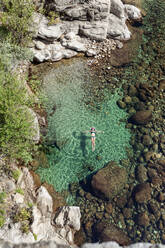 From above aerial view of slim female in swimwear swimming in transparent water near stony shore on resort - ADSF13782