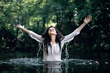 Cheerful female in transparent wet dress standing in pond water with closed eyes and outstretched arms - ADSF13790