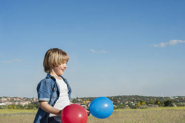 Side view of content kid standing with colorful air balloons in meadow and looking away while enjoying sunny day - ADSF13794