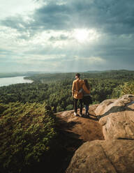 Rückenansicht eines anonymen Paares von Reisenden, die auf einem steinigen Pfad zwischen grünen Pflanzen in der Nähe eines Sees stehen und die spektakuläre Landschaft an einem sonnigen Tag genießen, während sie den Algonquin Provincial Park in Ontario in Kanada besuchen - ADSF13802