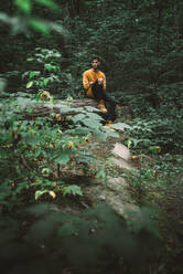 Junger männlicher Reisender, der auf einem Baumstamm im dichten grünen Laub sitzt und wegschaut, während er sich bei einer Wanderung im Wald im Algonquin Provincial Park in Kanada ausruht - ADSF13806
