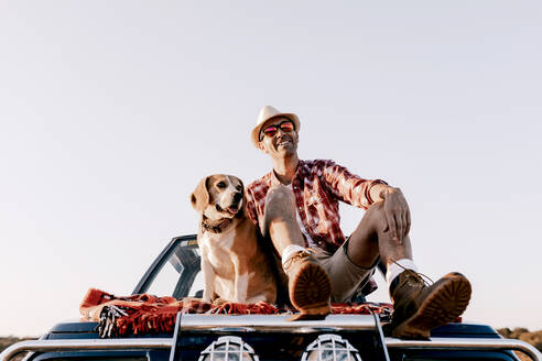 Verträumter männlicher Reisender mit Hut und Sonnenbrille, der mit herausgestreckter Zunge auf einem Auto in der Nähe eines reinrassigen Hundes sitzt und den Fluss und die Berge an einem sonnigen Tag bewundert - ADSF13809