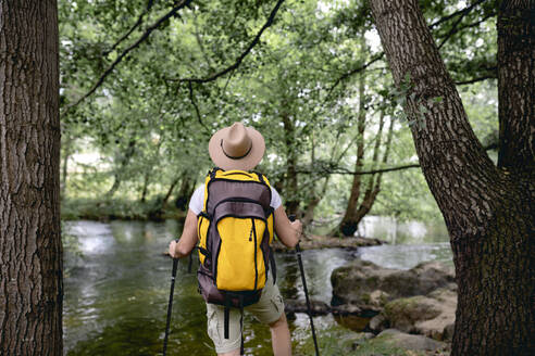 Junger Mann auf einem Wanderweg mit seinem gelben Rucksack und Hut auf dem Kopf an einem See mit vielen Bäumen und Naturgebieten mit Blick auf die Landschaft - ADSF13812