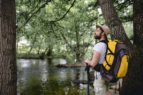 Junger Mann auf einem Wanderweg mit seinem gelben Rucksack und Hut auf dem Kopf an einem See mit vielen Bäumen und Naturgebieten mit Blick auf die Landschaft - ADSF13813