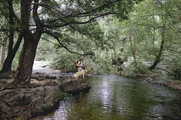 Landschaft eines jungen Mannes mit Hut und gelbem Rucksack, der mit seinem Hund über einen See mit Wasser und vielen grünblättrigen Bäumen wandert - ADSF13818