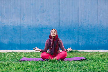 Gelassene arabische Frau in Sportkleidung und Hijab, die mit geschlossenen Augen im Park auf einer Matte im Lotussitz sitzt und mit geschlossenen Augen Yoga in Padmasana macht - ADSF13822