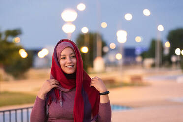 Erfreute ethnische Frau, die abends auf der Straße steht und den Hijab anlegt, während sie wegschaut - ADSF13827