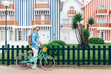 Seitenansicht eines trendigen Touristen mit Vintage-Fahrrad und Rucksack vor den hellen Gebäuden der Costa Nova du Prado - ADSF13832