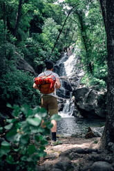 Rückenansicht eines nicht erkennbaren männlichen Reisenden mit Rucksack, der auf einem Felsen steht und die herrliche Landschaft eines grünen Waldes mit einem schnellen Fluss bewundert - ADSF13837