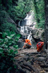 Seitenansicht des Reisenden mit Rucksack sitzt auf einem Felsen und bewundert erstaunliche Landschaft der grünen Wälder mit schnellen Fluss - ADSF13838