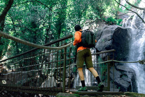 Sorgloser männlicher Tourist mit Rucksack, der entlang einer Hängebrücke aus Metall spaziert und die herrliche Natur des Waldes bewundert - ADSF13840