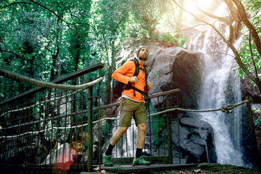 Sorgloser männlicher Tourist mit Rucksack, der entlang einer Hängebrücke aus Metall spaziert und die herrliche Natur des Waldes bewundert - ADSF13841