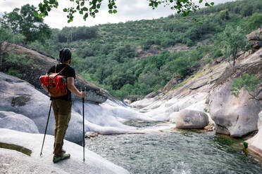 Rückenansicht eines anonymen männlichen Reisenden mit Rucksack und Trekkingstöcken, der auf einem Felsen steht und die malerische Aussicht auf einen Fluss im Hochland genießt - ADSF13846