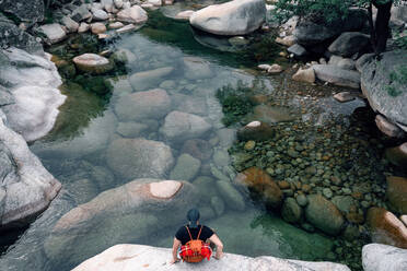 Anonymer männlicher Reisender mit Rucksack, der auf einem Felsen sitzt und die malerische Aussicht auf einen Fluss im Hochland genießt, von oben - ADSF13847