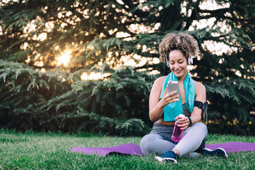 Fröhliche junge lockige Frau in Sportkleidung mit Kopfhörern sitzt im Gras und surft auf dem Smartphone, während sie sich mit einer Flasche Wasser nach dem Training im Sommerpark ausruht - ADSF13848